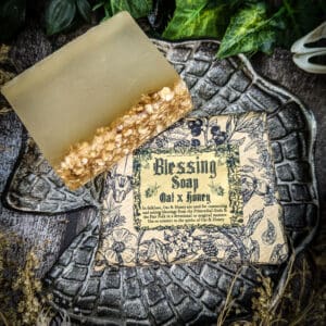 A handmade bar of Blessing Soap rests on a dark textured dish. The soap has a golden hue with oat and honey pieces embedded on top. Its vintage-style label reads 'Blessing Soap – Oat & Honey,' surrounded by floral illustrations. Green leaves and dried flowers frame the scene.