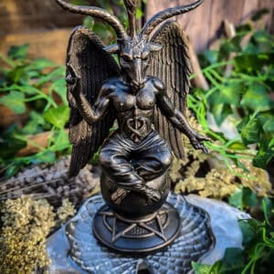 A bronze-colored statue of the Witch Lord, also known as Baphomet, sits cross-legged on a lotus pedestal with a pentacle base. The figure has goat horns, wings, and a torch above its head, surrounded by greenery and stone in an outdoor setting.