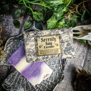 A bar of Serenity Soap in lavender rests on a dark floral-textured dish. The soap has a soft purple and white marbled design, placed beside its vintage-style illustrated label. Green leaves and rustic wooden background frame the scene