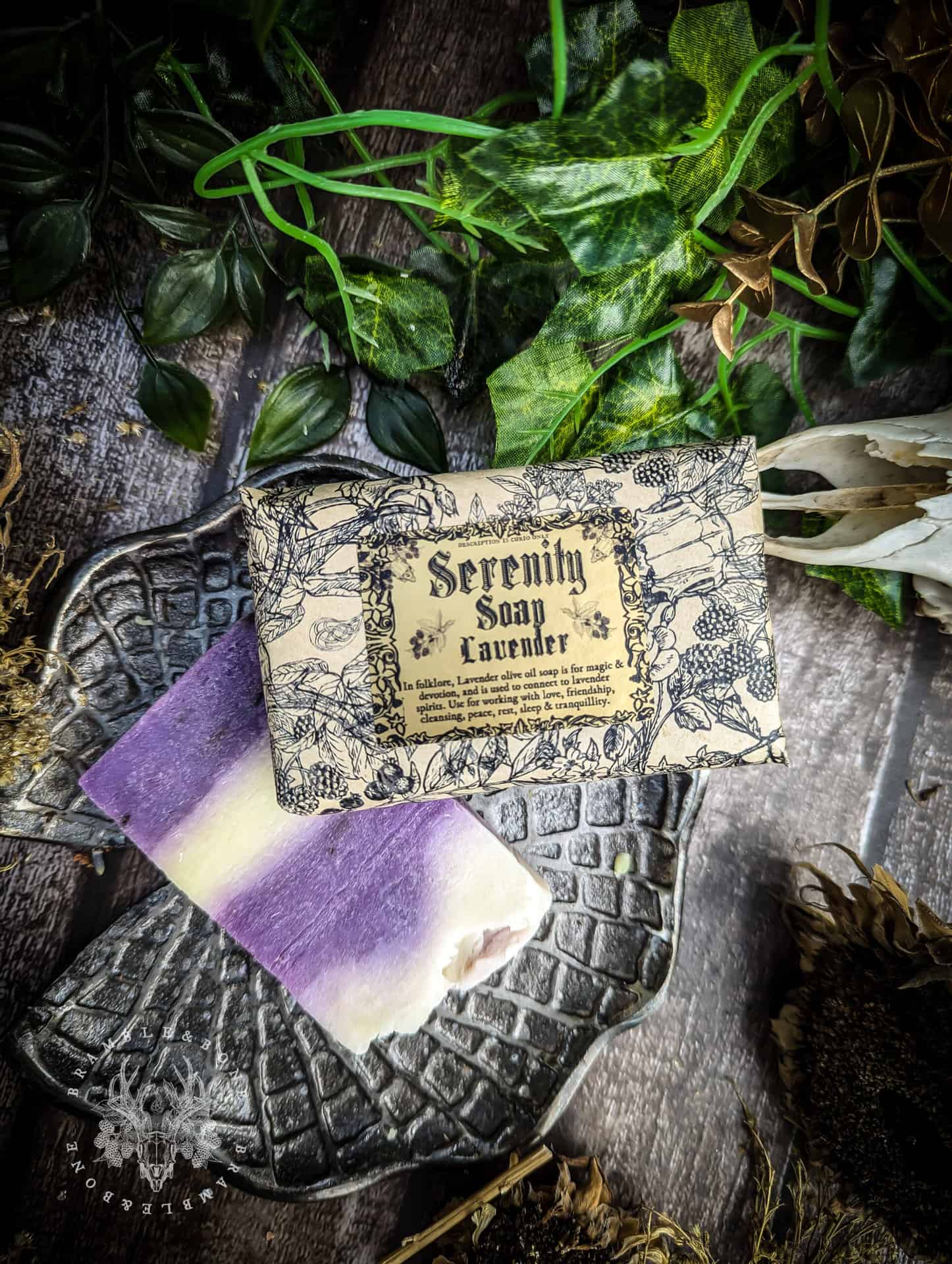A bar of Serenity Soap in lavender rests on a dark floral-textured dish. The soap has a soft purple and white marbled design, placed beside its vintage-style illustrated label. Green leaves and rustic wooden background frame the scene