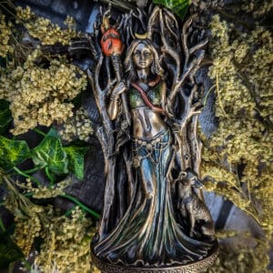 Hekate and Hound statue showing the Greek goddess of witchcraft standing before a tree, holding a torch, with her loyal hound at her side, detailed in bronze and green tones, surrounded by ivy and dried flowers on a rustic backdrop.