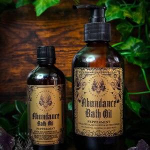 Amber bottles of Abundance Bath Oil on dark wood, ornate label, surrounded by polished amethyst and green vines.