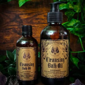 Amber glass bottles labeled “Cleansing Bath Oil” on a dark wooden surface with an ornate gothic beige label. Styled with trailing green ivy and amethyst crystals to suggest purification and renewal.