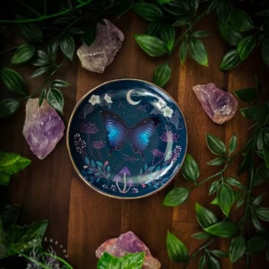 A decorative moth trinket dish featuring a luminous blue-purple butterfly on dark teal background, surrounded by moths, mushrooms, crescent moon, ivy, and wildflowers with gold rim. The dish sits on weathered wood, framed by trailing green foliage and raw amethyst crystals, creating an enchanted folk magic aesthetic perfect for altar work and fairy faith practice.