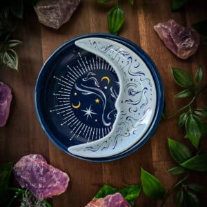 Circular ceramic trinket dish featuring a crescent moon design with celestial night sky imagery displayed on weathered wood. The dish shows a pale silver moon face surrounded by stars and crescents on a deep blue-black centre, photographed with purple amethyst crystals and green foliage creating an altar-like atmosphere.