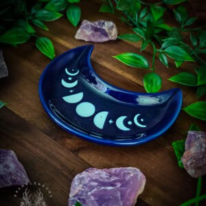 A deep navy blue crescent moon ceramic dish with mint-painted moon phase markings sits on dark wood, surrounded by rough raw amethyst crystals and lush green foliage. The full lunar sequence - new moon to full and back - runs visibly along the dish's inner curve. The Bramble and Bone logo sits in the lower left. Natural lighting creates a rich, atmospheric composition.