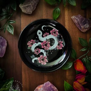 Black trinket dish featuring white serpent design holds a white tealight candle, surrounded by dried rose petals in deep pink and golden orange, purple amethyst crystals, and small dried flowers. Set on dark wooden surface with soft green foliage visible in background. The serpent curls among pink flowers against a starry night sky.