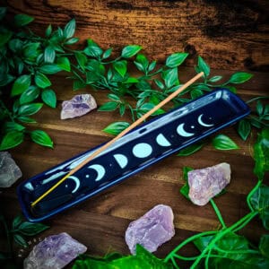 Deep navy glazed ceramic incense ash catcher lying diagonally on dark wood planking, a single wooden incense stick resting across its length. The full lunar cycle is depicted in white along the surface, from thin crescent through full moon and back again. Raw amethyst clusters and trailing green foliage surround the piece. Bramble & Bone watermark in the lower left corner.