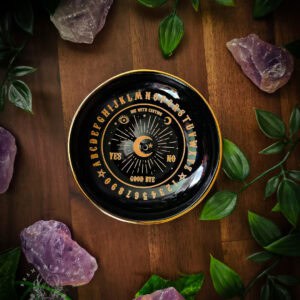 Black ceramic talking board incense holder on weathered wood, framed by raw amethyst clusters and trailing green foliage. Gold ouija-style lettering circles the dish - alphabet, numbers, YES, NO, GOOD BYE - with a radiant crescent moon at the centre. The text USE WITH CAUTION arches above the planchette motif. Warm light catches the premium gold finish against the dark glaze.