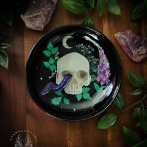 A glossy black ceramic trinket dish featuring a pale skull with a purple serpent coiling through its eye socket, surrounded by green ivy, purple foxglove flowers, and a crescent moon against a starlit background. The dish sits on dark wood, framed by raw amethyst crystals and trailing greenery, evoking traditional witchcraft altar aesthetics.