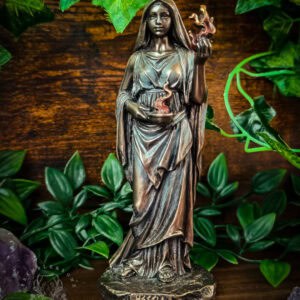 Bronze-finish Hestia statue standing in flowing robes with one hand raised holding a flame and the other cradling a bowl, photographed against weathered wood surrounded by vibrant green foliage. The polyresin goddess figure displays intricate detailing in the draped fabric and serene facial features, with 'HESTIA' inscribed on the base. Flanking crystals and lush plant life create an altar-like composition.