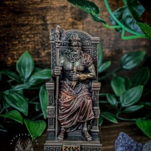 Zeus mini statue in hand-painted bronze seated on ornate throne. Front-facing view shows detailed Greek key patterns on base spelling 'ZEUS', decorative throne with eagle perched above, flowing robes and beard. Surrounded by green foliage and blue crystal on wooden surface. Warm metallic tones throughout.